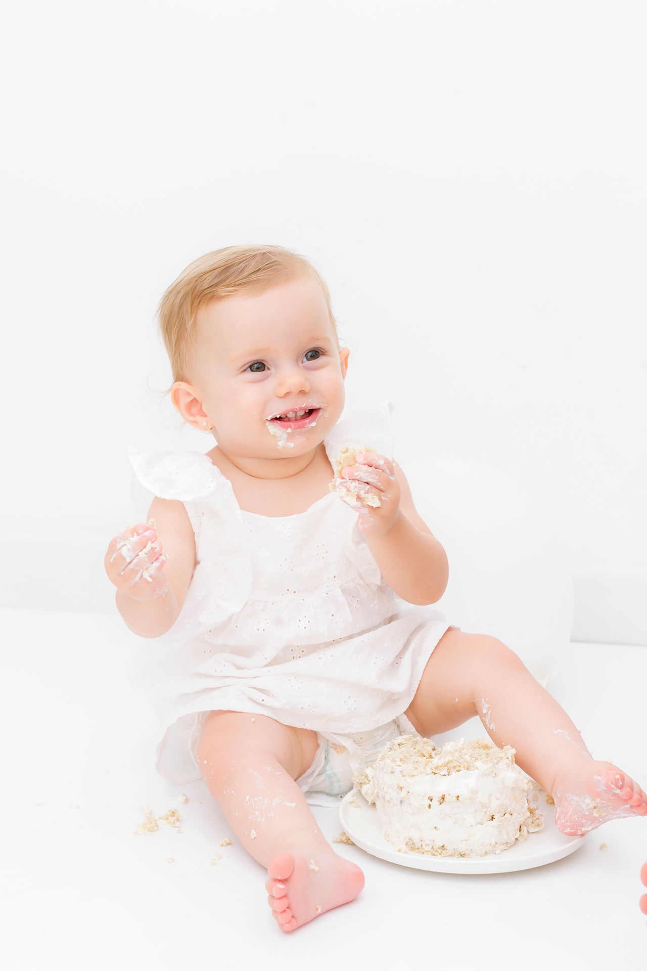 foto de un bebe de un año festejando su cumpleaños con un pastel con cubierta blanca, vestido blanco, fondo blanco