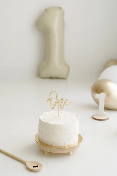 Tarta smash cake blanca junto a una cuchara de madera, un número uno pequeño y un fondo decorado con globos blancos, dorados y un globo gigante verde claro con la forma del número 1.