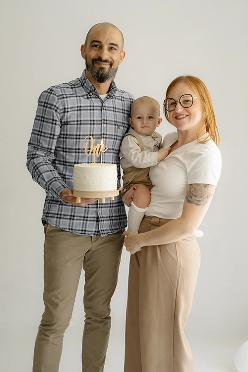 Padres de pie sonriendo a la cámara y sosteniendo en brazos a su bebé junto a su pastel de primer cumpleaños en una sesión smash cake.
