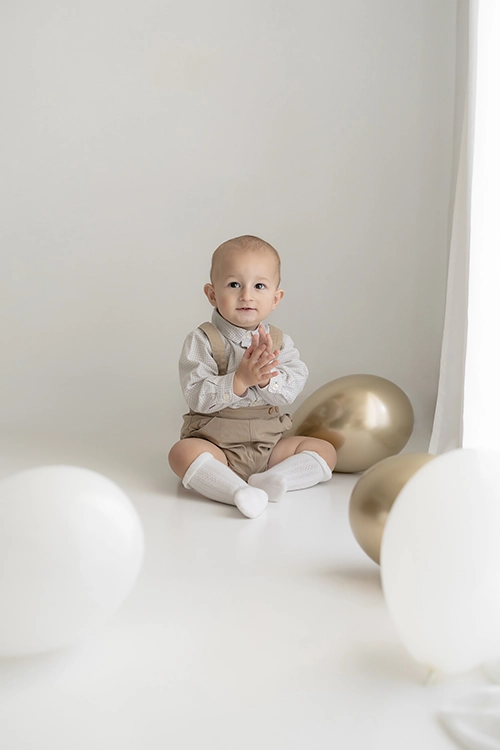 Niño pequeño sentado hacia la cámara en un decorado de cumpleaños con globos blancos, dorados en sesion de fotos smash cake.