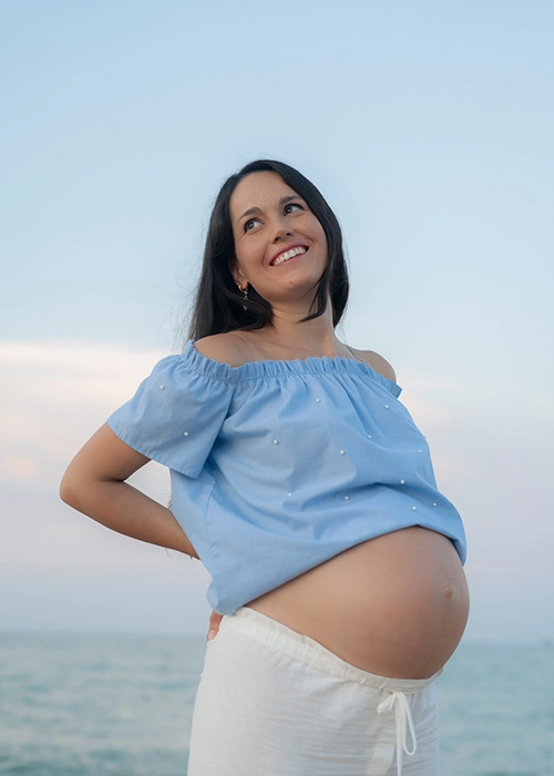 mujer embarazada sonriente en la playa con blusa azul claro y pantalón beige