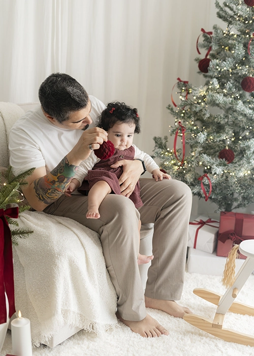 padre junto a su hija bebe sentada en sus piernas escenario de navidad blanco y burdeos