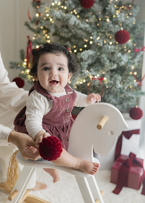 niña bebe mirando a cámara en caballito de madera balancín sonriendo en escenario de navidad