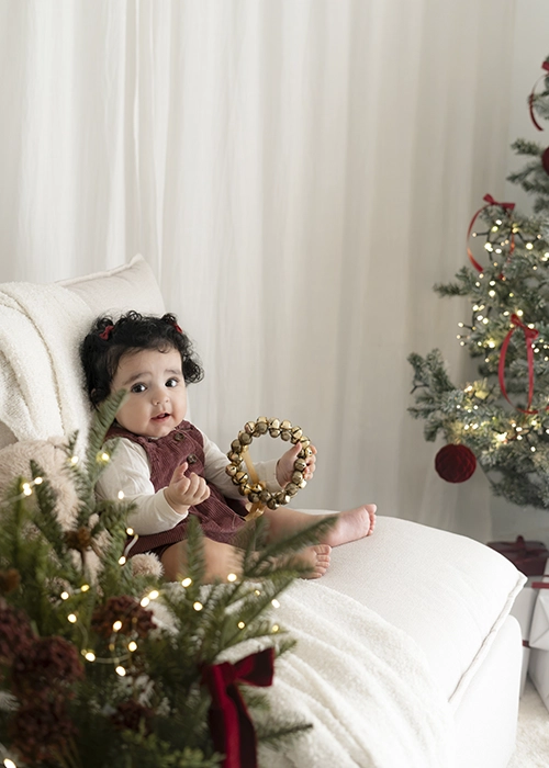 niña de un año sentada en sofá al lado de árbol de navidad con luces