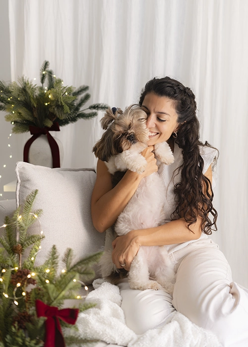 mujer con su perrito pequeño en brazos abrazándolo en escenario de navidad blanco y burdeos